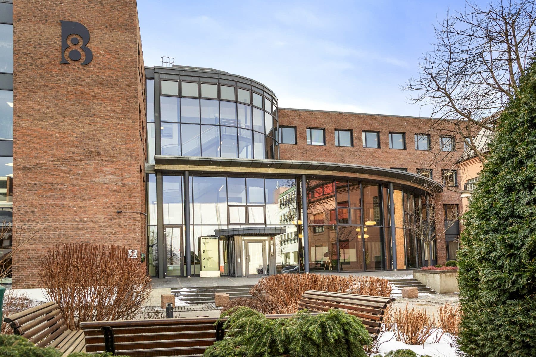 Inngangsparti i glass mellom teglfasader, med benker og beplantning foran